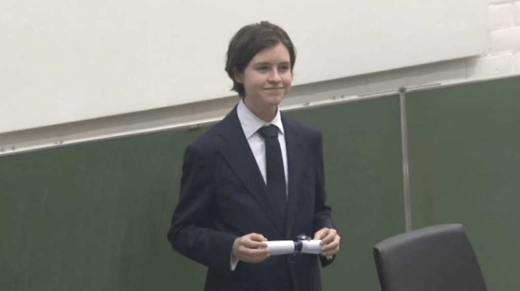 一名少年穿著西裝，微笑著手持博士學位証書，背景為課堂環境。