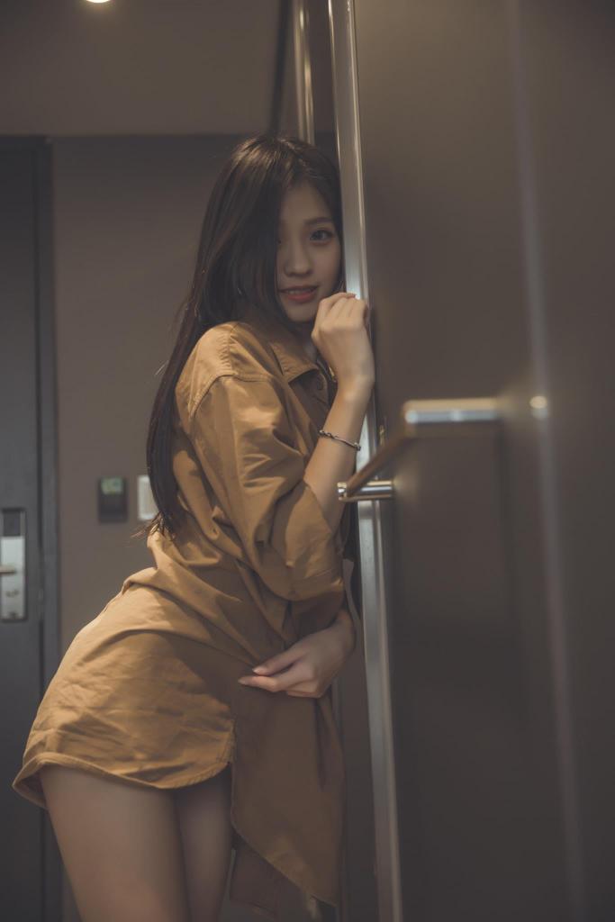 一位年輕女性穿著棕色上衣，微笑著靠在門邊，背景是室內環境。