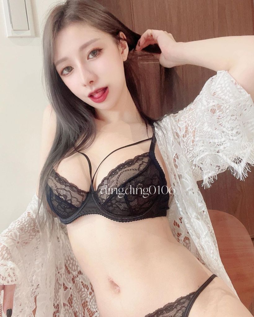一名女性模特穿著黑色蕾絲內衣，搭配白色蕾絲外套，擺出時尚姿勢，坐在室內背景中。