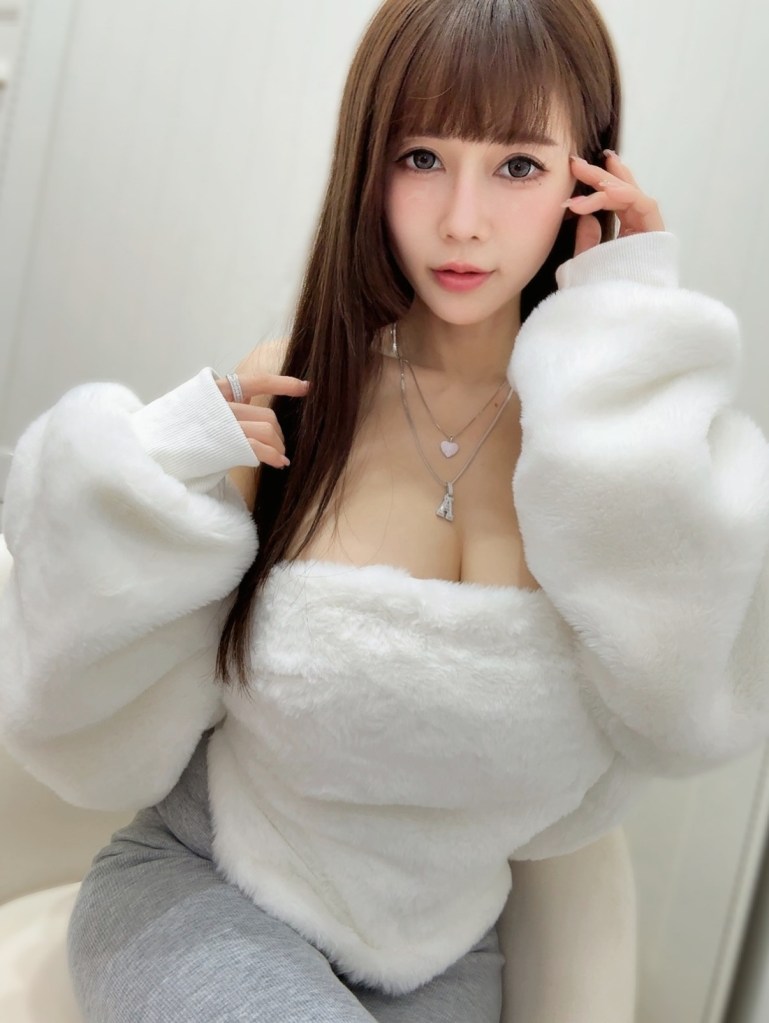 一位女性坐在椅子上，穿著白色毛衣和灰色緊身褲，長髮披肩，手指輕撫臉頰，背景模糊。