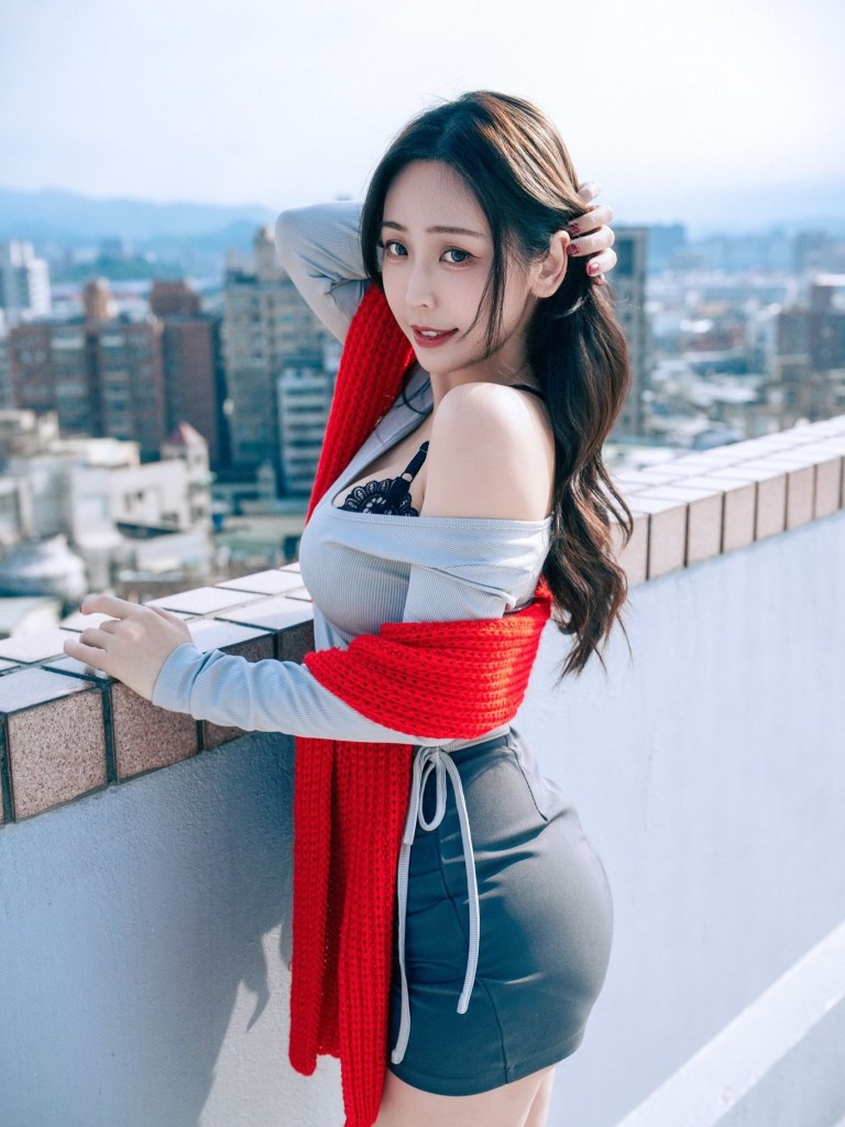 一位女性站在城市高樓的邊緣，穿著灰色上衣和黑色裙子，搭配紅色圍巾，背景是城市景觀和藍天。