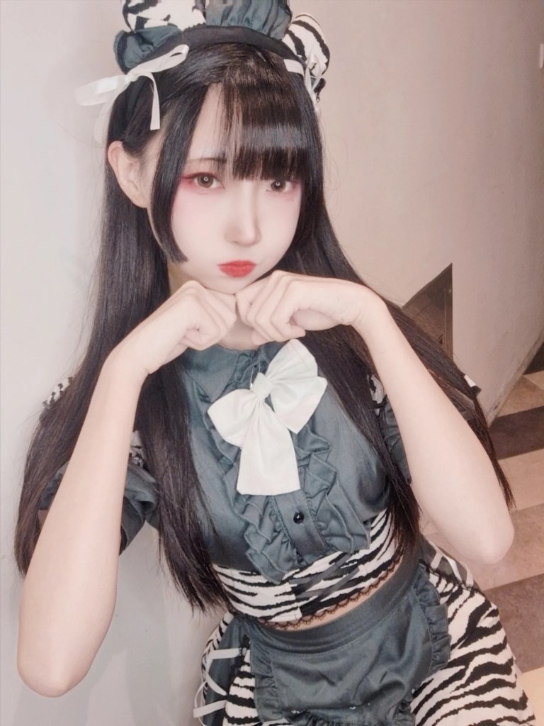 一位穿著黑白相間的服裝少女，微微撅嘴並用手握拳擺出可愛姿勢，背景為簡單的牆面。