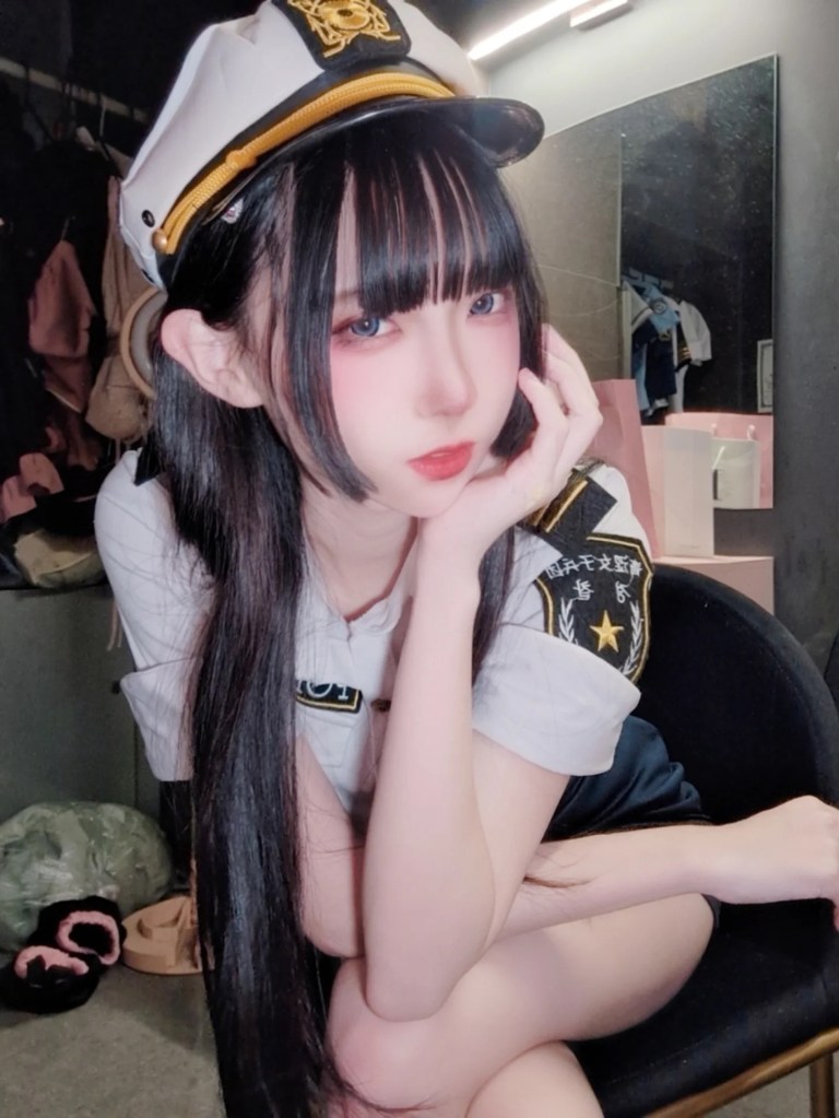 一位穿著制服的女性坐在椅子上，手托著下巴，展現出沉思的神情，背景中可見衣物和其他物品。