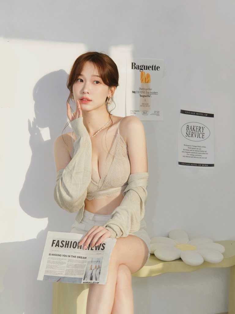 一位女性坐在椅子上，穿著淺色內衣和開襟毛衣，手中握著一份時尚報紙，背景有麵包和烘焙服務的海報，展現輕鬆的室內氛圍。