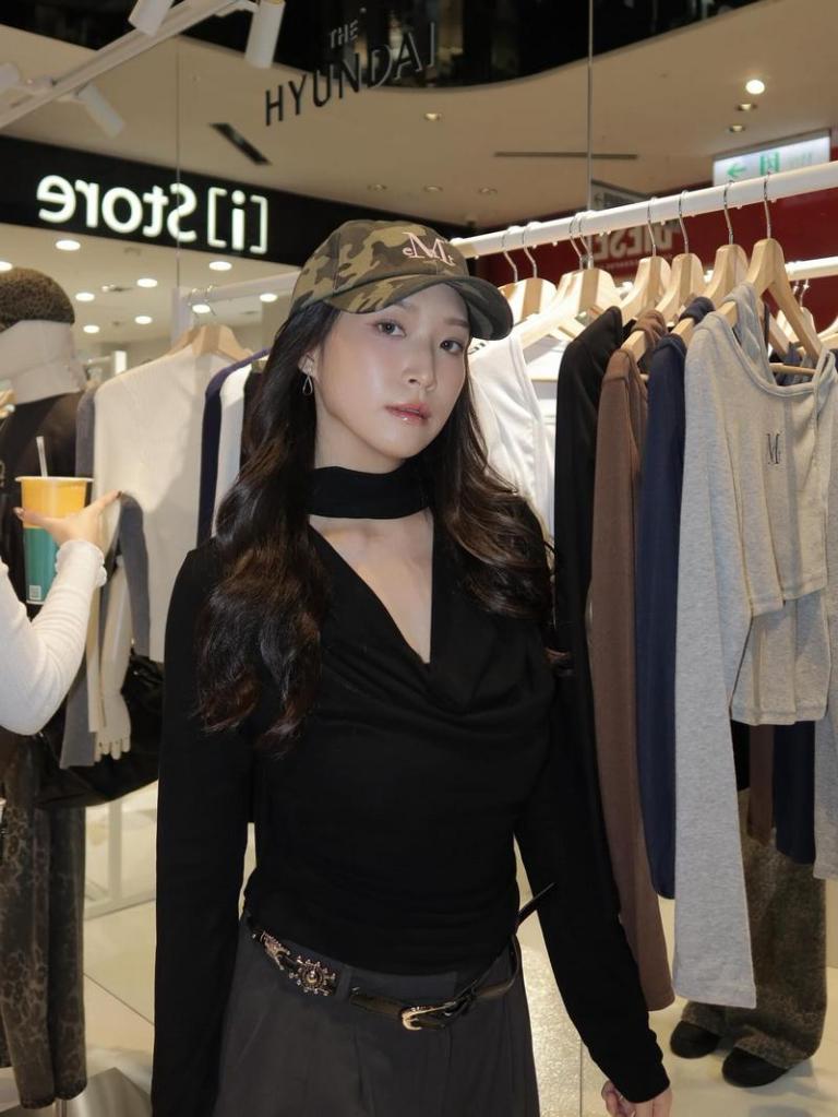 一位穿著黑色上衣和帽子的女性在商店內部，背景有衣物架和購物者。