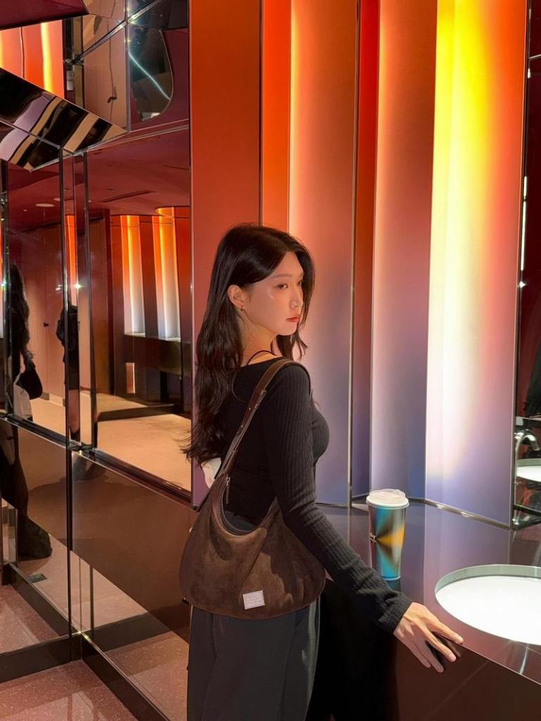 一位女性站在色彩斑斕的鏡面牆前，穿著黑色長袖上衣，肩上背著一個深色手袋，手中拿著一杯飲品。