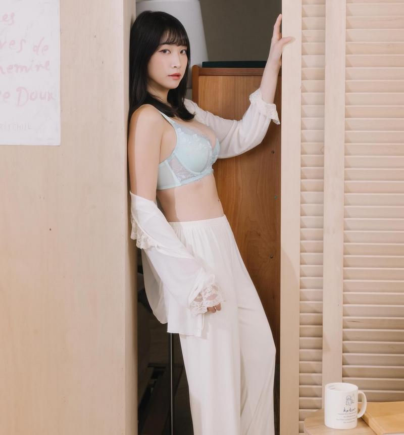 一位女性穿著淡藍色內衣和白色長褲，正倚靠在牆邊，展現自信的姿態。背景有木質家具和一杯白色咖啡杯。