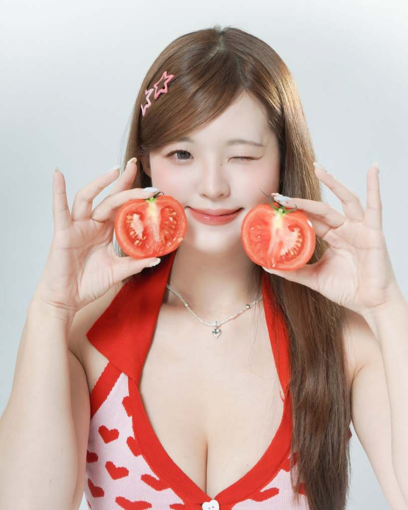 一位女士微笑並眨眼，手中各拿著一半的西紅柿，穿著紅色與心形圖案的上衣。