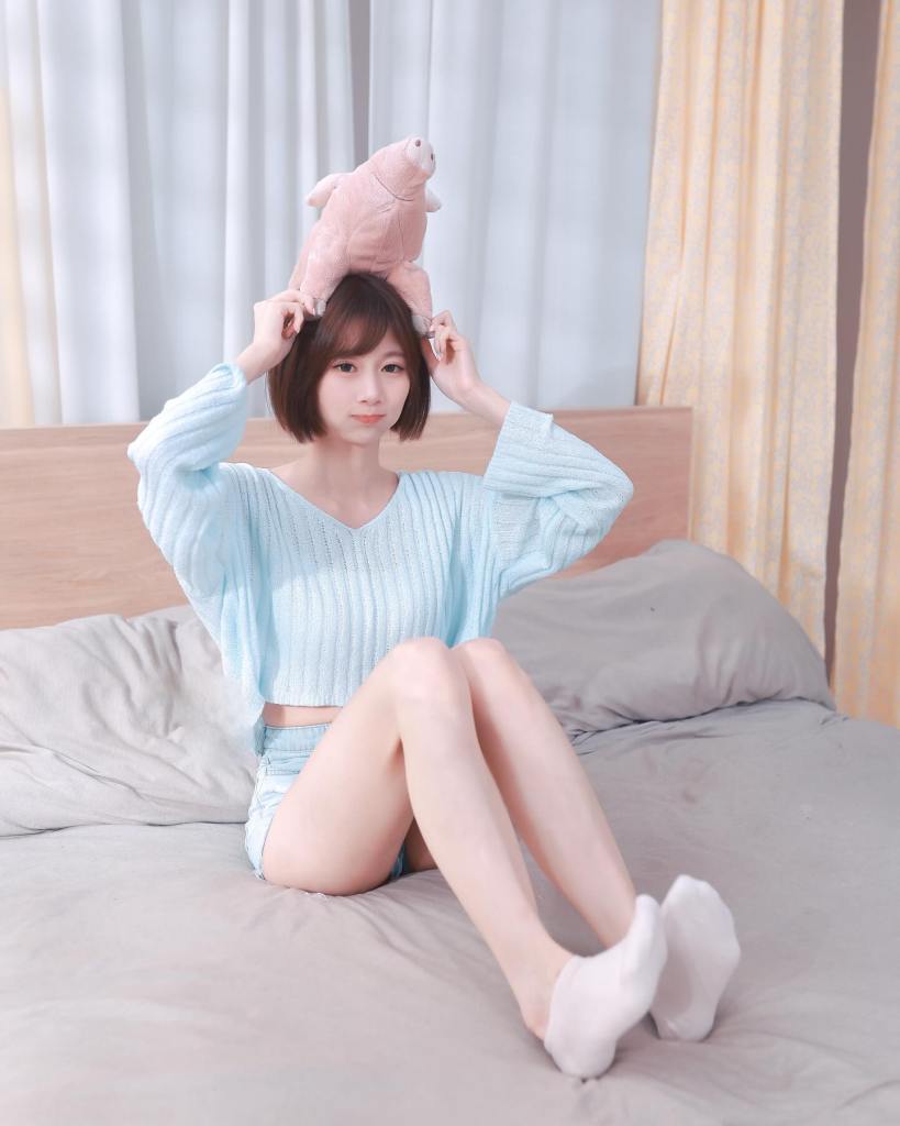 一名女孩坐在床上，頭上放著一隻粉紅色的豬形玩具，穿著淺藍色的針織上衣和短褲。