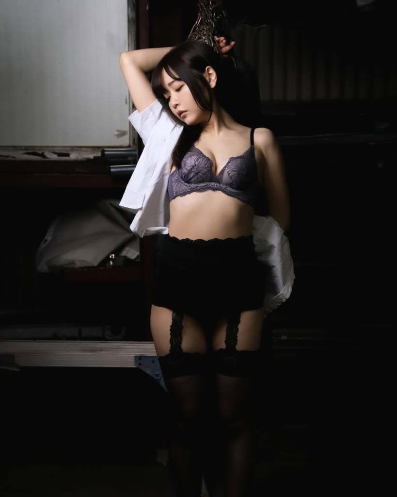 一位女性模特身穿蕾絲胸罩和黑色短褲，手臂高高舉起，面容優雅，站在昏暗的環境中。