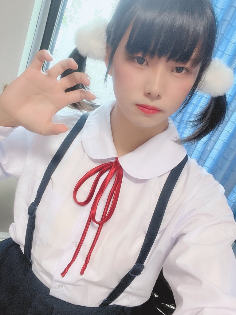 一位穿著白色襯衫和黑色吊帶裙的年輕女性，配有紅色絲帶，雙馬尾處有白色毛球，面對鏡頭作出可愛的手勢，背景為藍色窗簾