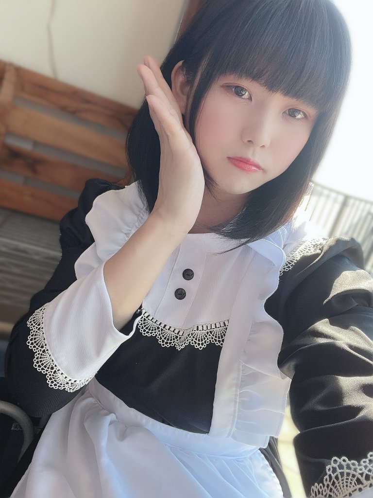 一位穿著黑白女僕服裝的年輕女性，面對鏡頭微笑，手放在臉旁，背景為戶外座位區