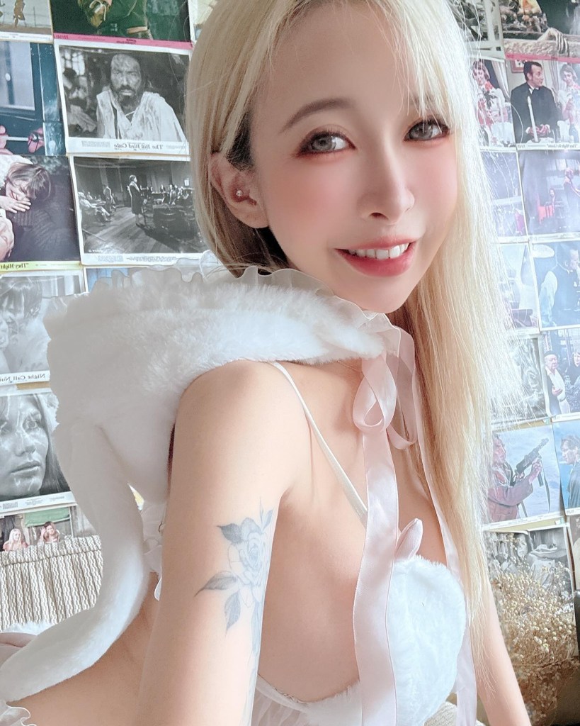 一位金髮女性微笑著，身穿白色衣物，背景是多幅黑白照片。