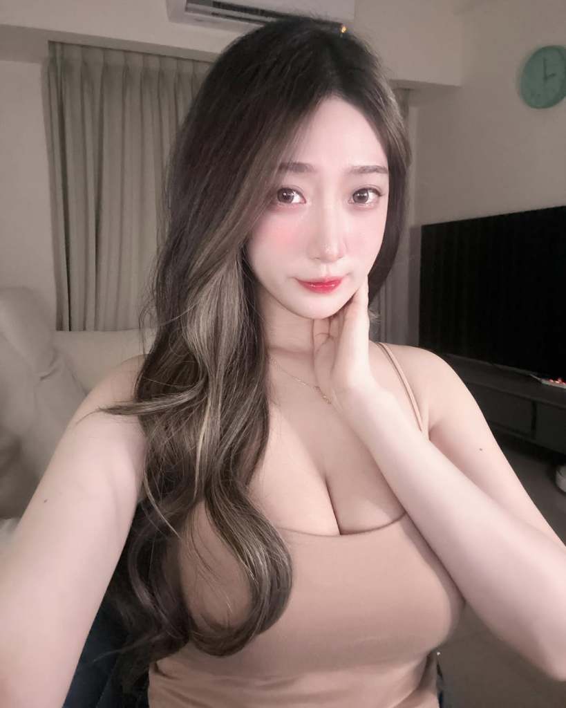 女性自拍，長髮披肩，穿著淺色上衣，背景為室內環境。