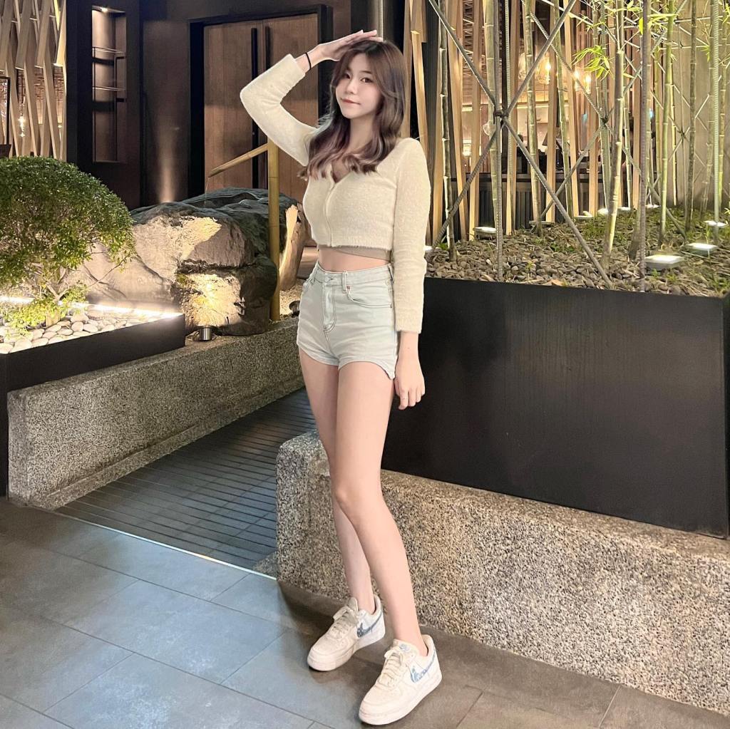 一名年輕女性在室外環境中站立，穿著淺色短褲和針織上衣，搭配白色運動鞋，手放在額頭上，背景有綠色植物和石頭裝飾。