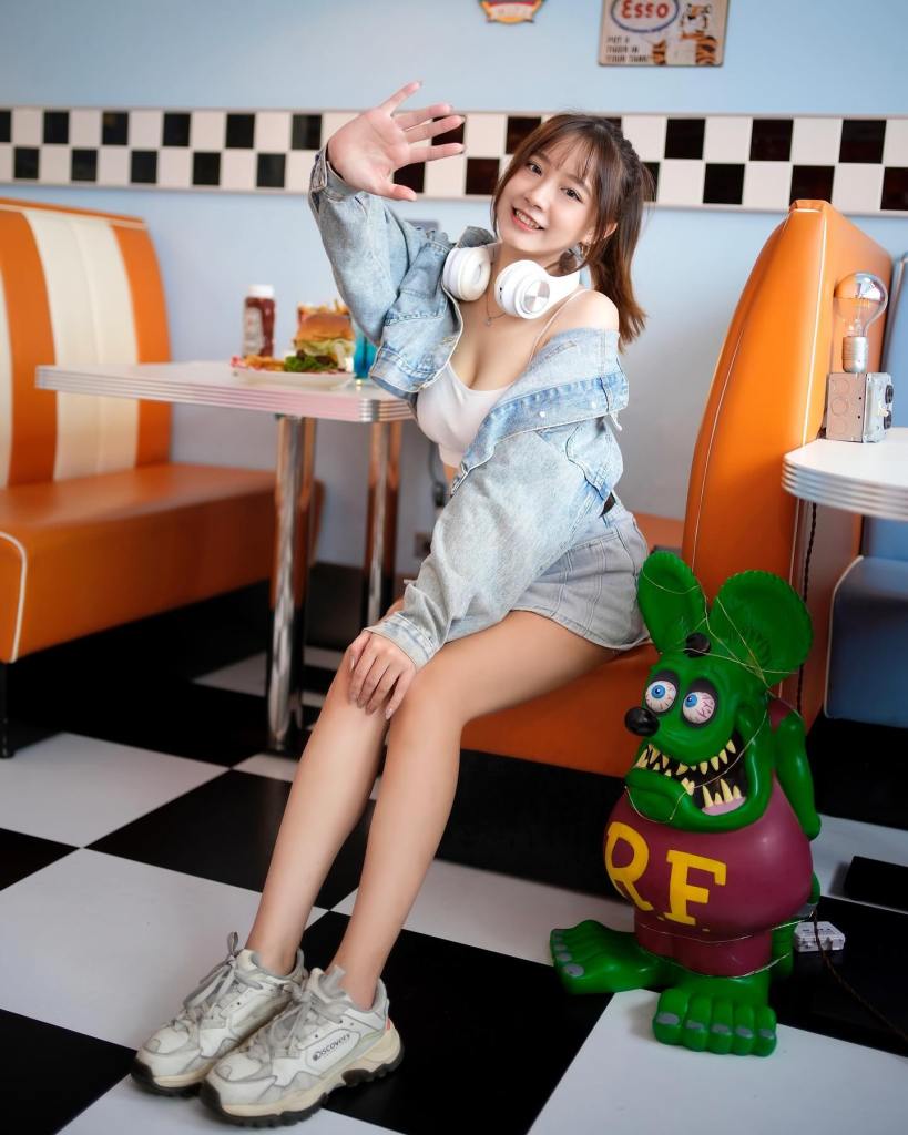 一名女性坐在餐廳內，穿著牛仔外套和耳機，微笑並揮手，旁邊有一個綠色的玩具角色，背景是彩色的座位和黑白棋盤地板。