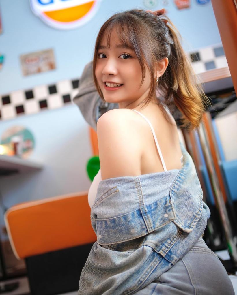 年輕女性微笑著，穿著露肩上衣和牛仔外套，背景是色彩豐富的餐廳環境