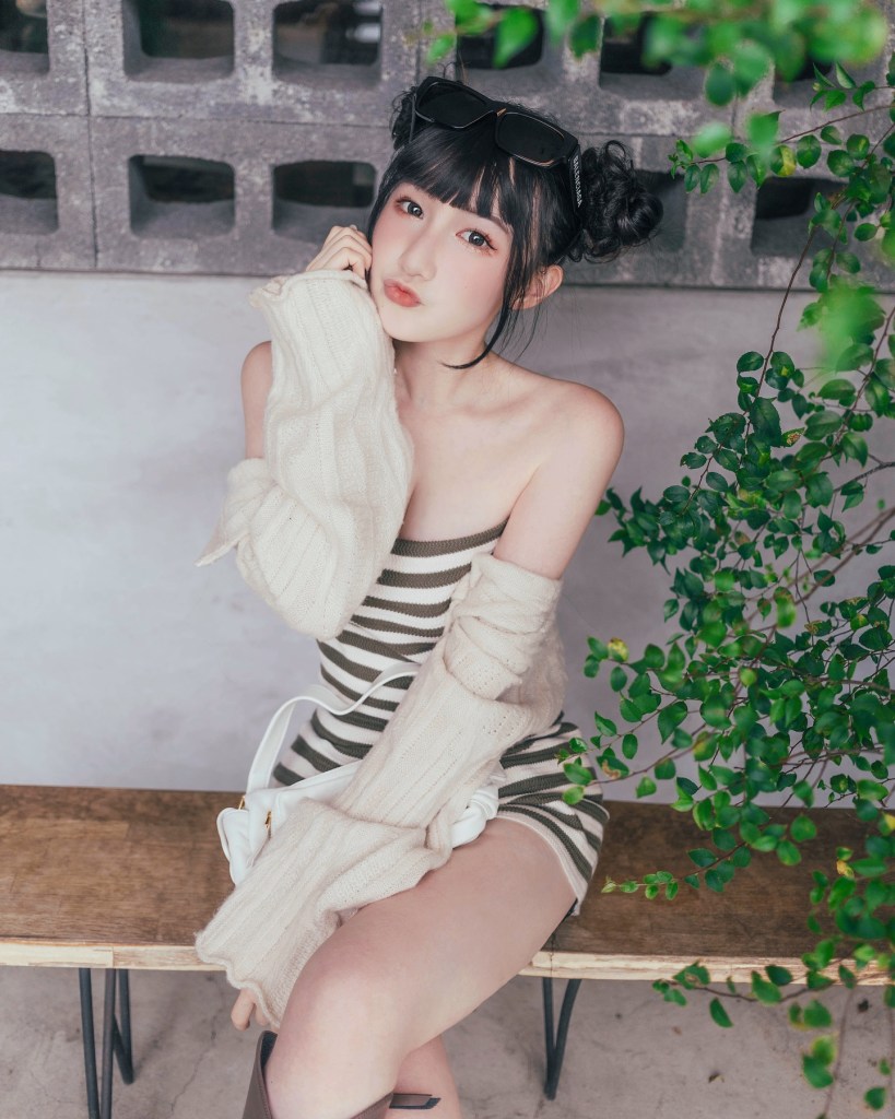 穿著條紋連衣裙的年輕女性，坐在長椅上，手中握著毛衣袖子，背景有綠色植物。