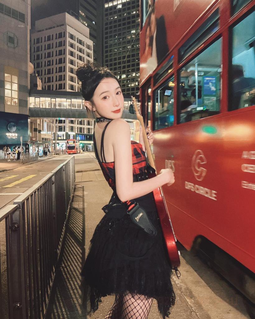 一位身穿紅黑格子裙子的女子，在夜晚的香港街頭，手持吉他，背景是紅色的雙層巴士。