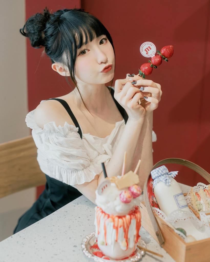 一位女孩穿著白色上衣，拿著三顆草莓，臉上露出撅嘴的表情，桌上有一杯冰淇淋和小籃子食物。