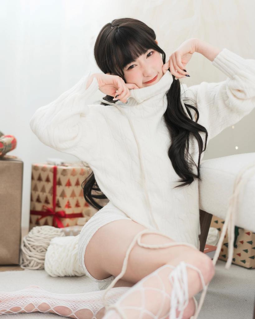 一名女性穿著白色毛衣，坐在地上，微笑著將毛衣的領口拉過下巴，背景有包裝好的禮物和裝飾品。