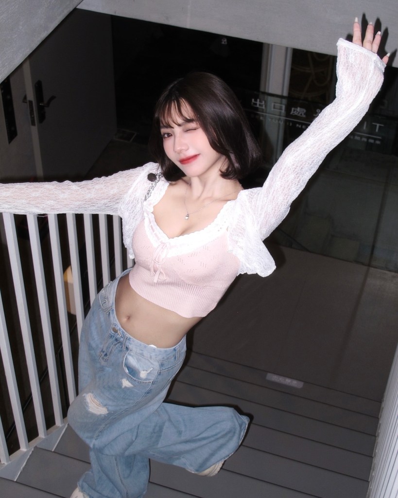 一位女孩在樓梯上擺姿勢，穿著粉色上衣和寬鬆的牛仔褲，微笑並眨眼，顯得活潑可愛。