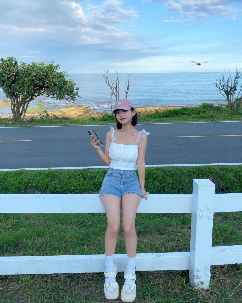 一位年輕女性穿著白色上衣和牛仔短褲，坐在白色圍欄旁，手中拿著手機，背景是海邊和藍天，遠處可見樹木和一隻飛翔的鳥。