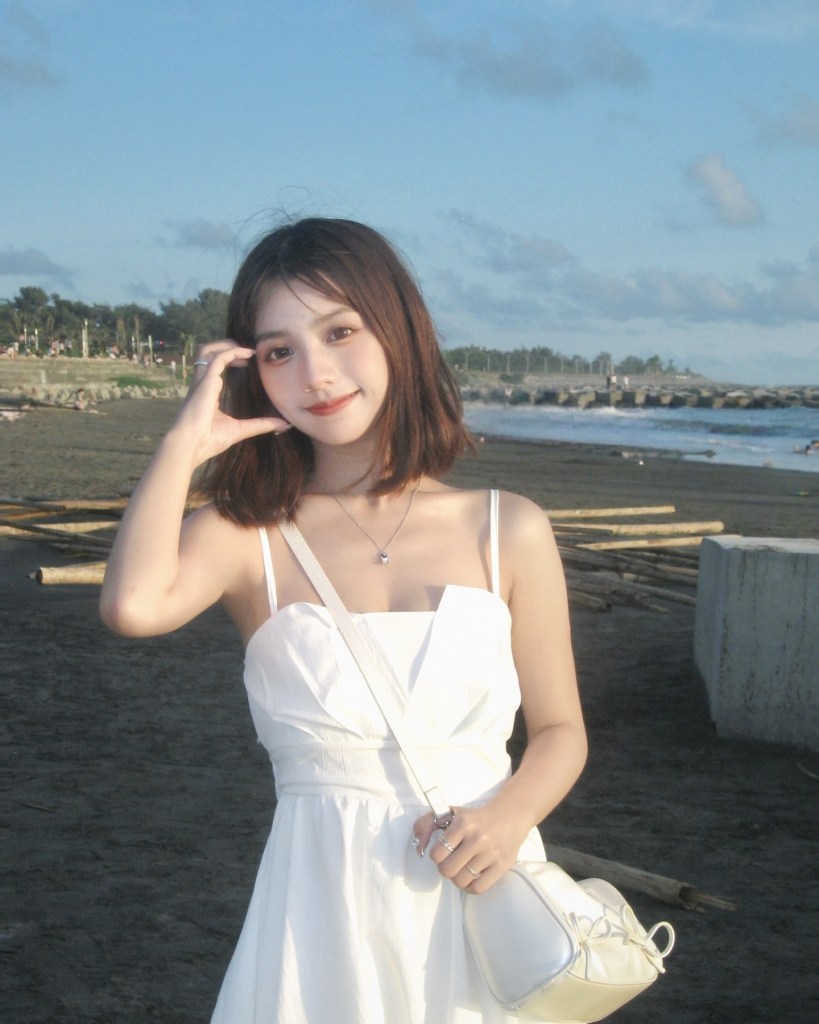 一位穿著白色連衣裙的年輕女性，站在海灘上，微笑著，手放在頭髮上，背景是藍天和沙灘。