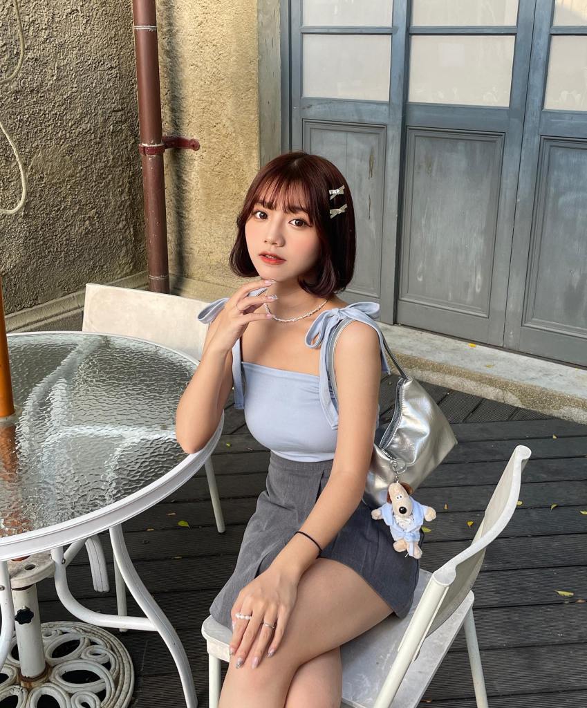 一位年輕女性坐在戶外桌子旁，穿著淺藍色上衣和灰色短裙，手中輕輕撫摸著手指，背著一個銀色手袋，手袋上有毛絨玩具掛飾。