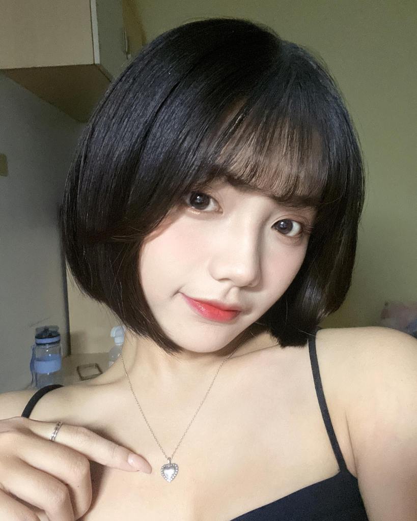 一位女性在室內拍攝自拍，穿著黑色背心，佩戴心形項鍊，表情自信微笑。