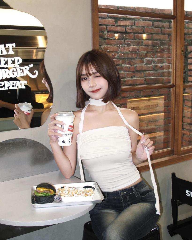 一位女性微笑著，手持飲料，與桌上食物一起拍照，背景有紅磚牆和鏡子。