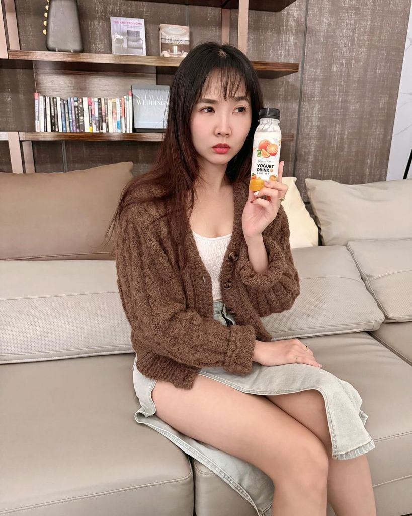 一位穿著棕色針織外套的女性坐在沙發上，手持一瓶酸奶飲品，神情凝重，背景是書架和柔軟的沙發。
