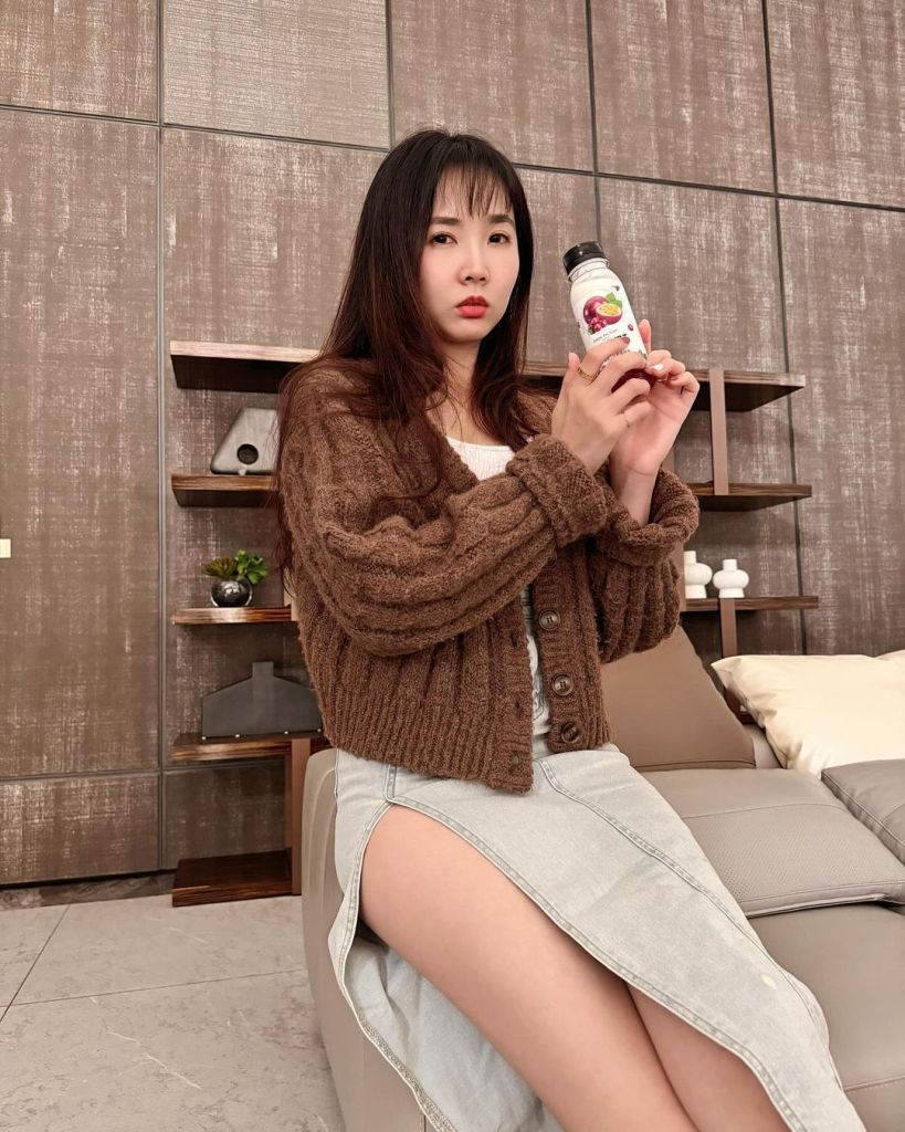 一位穿著棕色毛衣和牛仔裙的女性，手持飲料瓶，坐在沙發上。