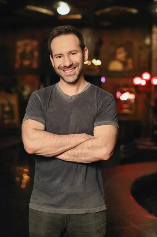 一名男性在室內微笑，手臂交叉，穿著深色T恤，背景模糊，燈光柔和。