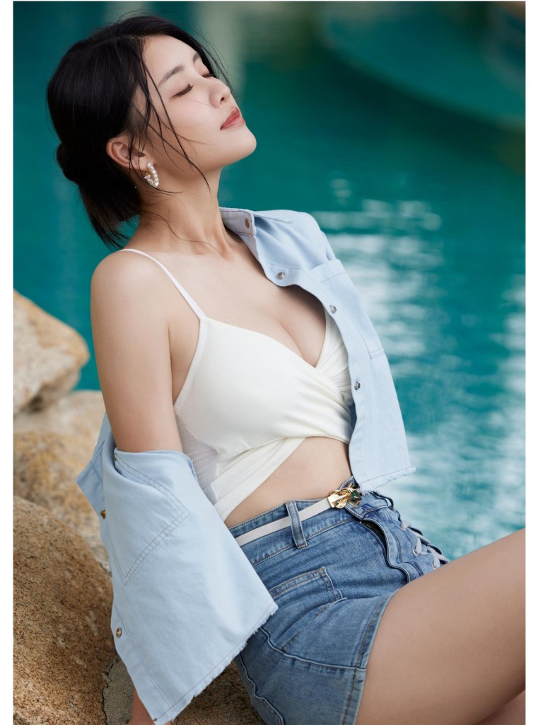 一位女性坐在水邊，穿著淺藍色外套和白色背心，面帶微笑，享受陽光，頭髮輕輕披肩。