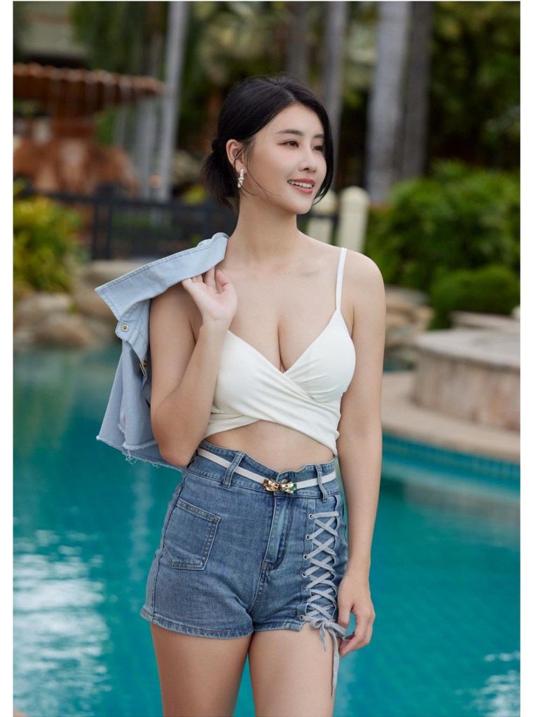 一位年輕女性穿著白色背心和牛仔短褲，站在泳池旁微笑，手中拿著一件外套，背景為綠色植物和泳池。