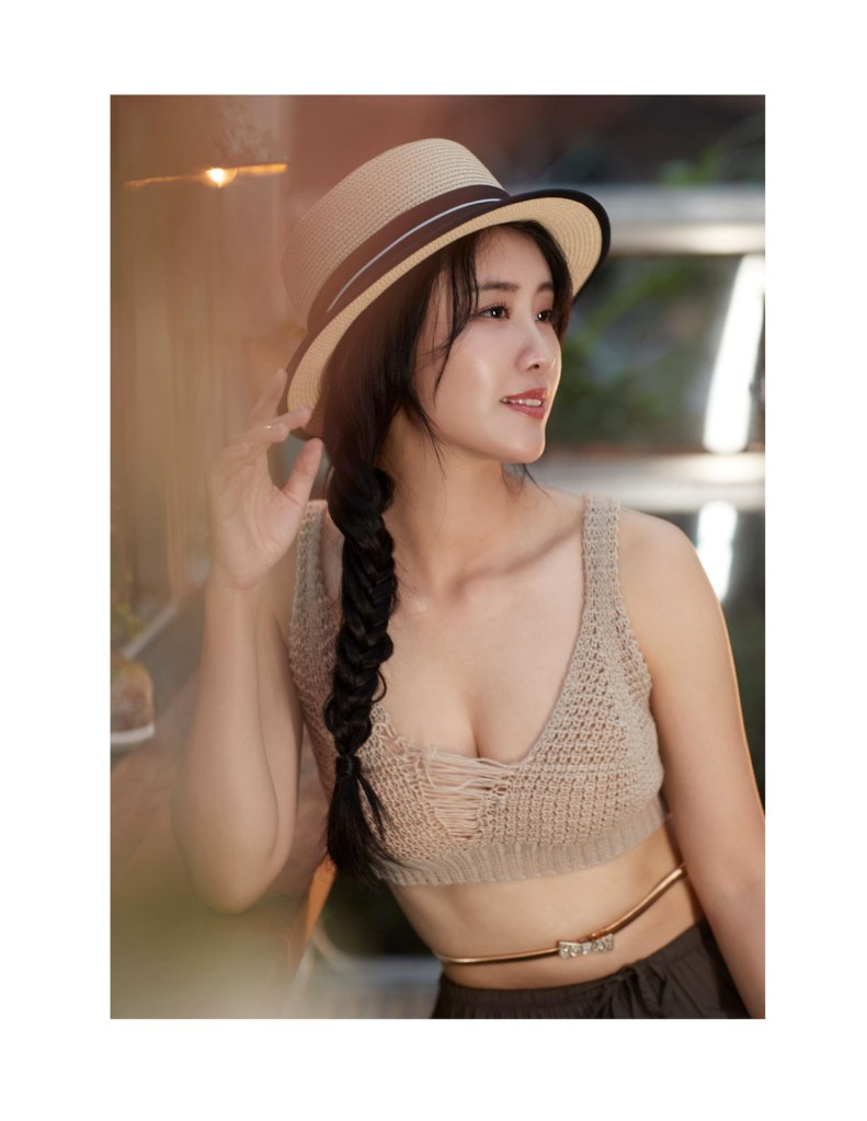 一位女性在戶外微笑，穿著編織的淺色上衣和黑色褲子，搭配草帽，披著辮子，背景模糊，散發著自然氛圍。