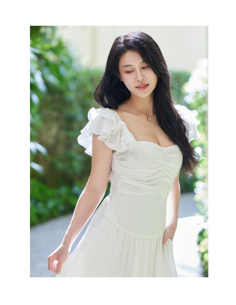 一位穿著白色裙子的女性，坐在綠色背景前，神情柔和，展現優雅和自信。