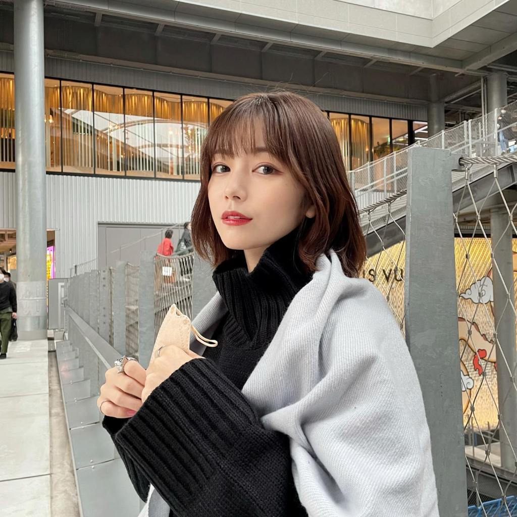 一位穿著黑色高領毛衣和灰色披肩的年輕女性，在現代建築背景下微笑，手中握著一個口罩。