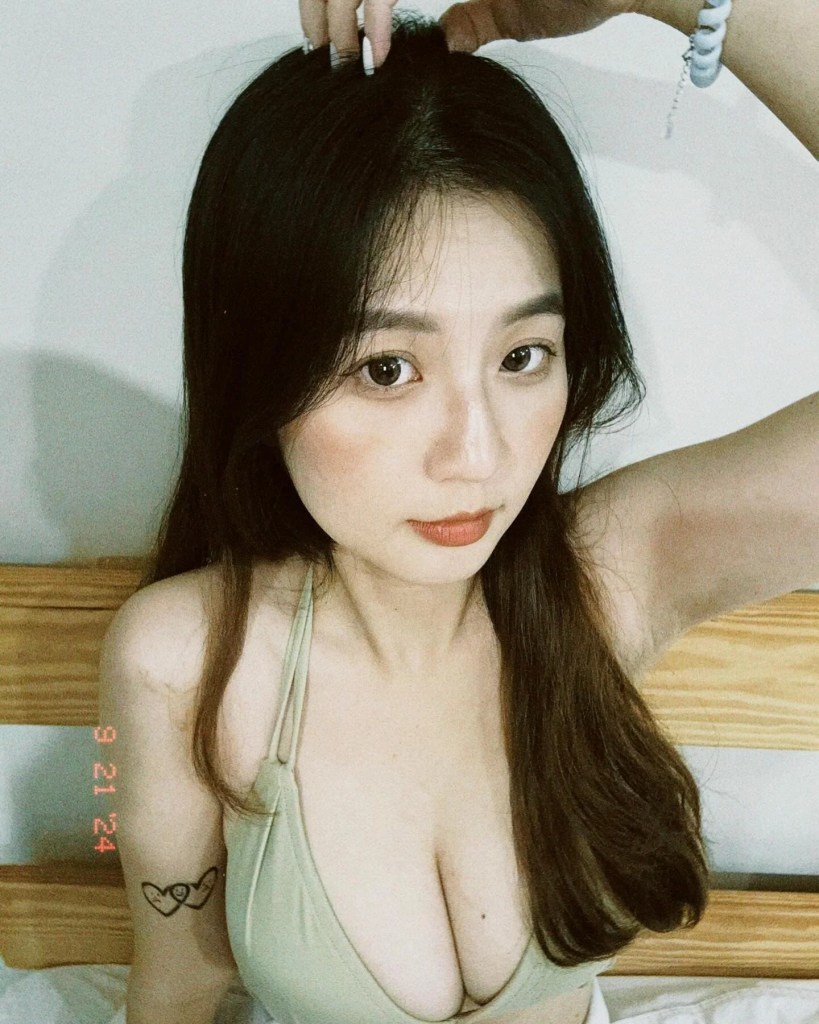 一位女性坐在床上，穿著淡綠色泳裝，上半身朝向鏡頭，手撥弄著頭髮，背景為淺色牆壁。