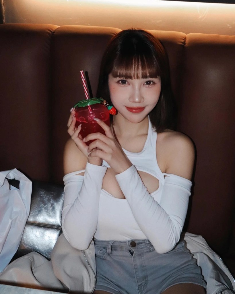 一位年輕女性坐在沙發上，手持一杯裝飾有綠色葉子和草莓的紅色飲品，微笑著看著鏡頭。