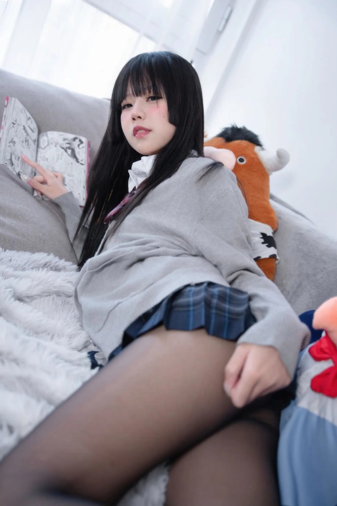 一位年輕女性坐在沙發上，穿著灰色毛衣和格紋裙子，手中翻閱漫畫。背景有一個毛茸茸的毯子和幾個玩具。