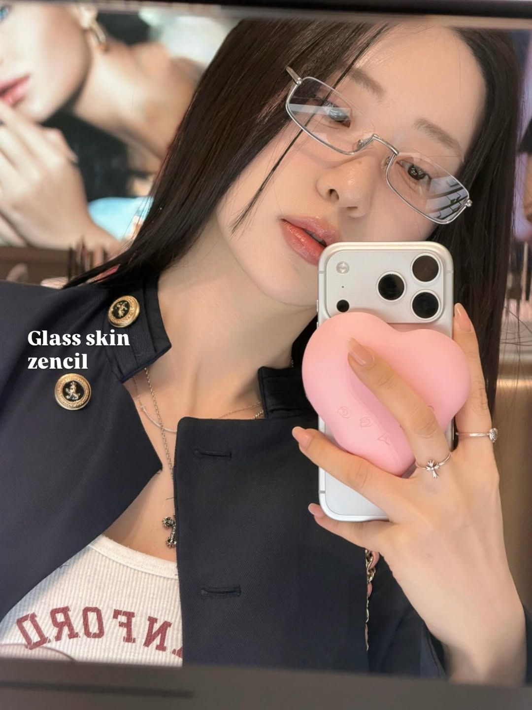 一名女性穿著黑色外套，手持手機自拍，臉上妝容光滑，佩戴眼鏡，背景中展現了另一位女性的影像。