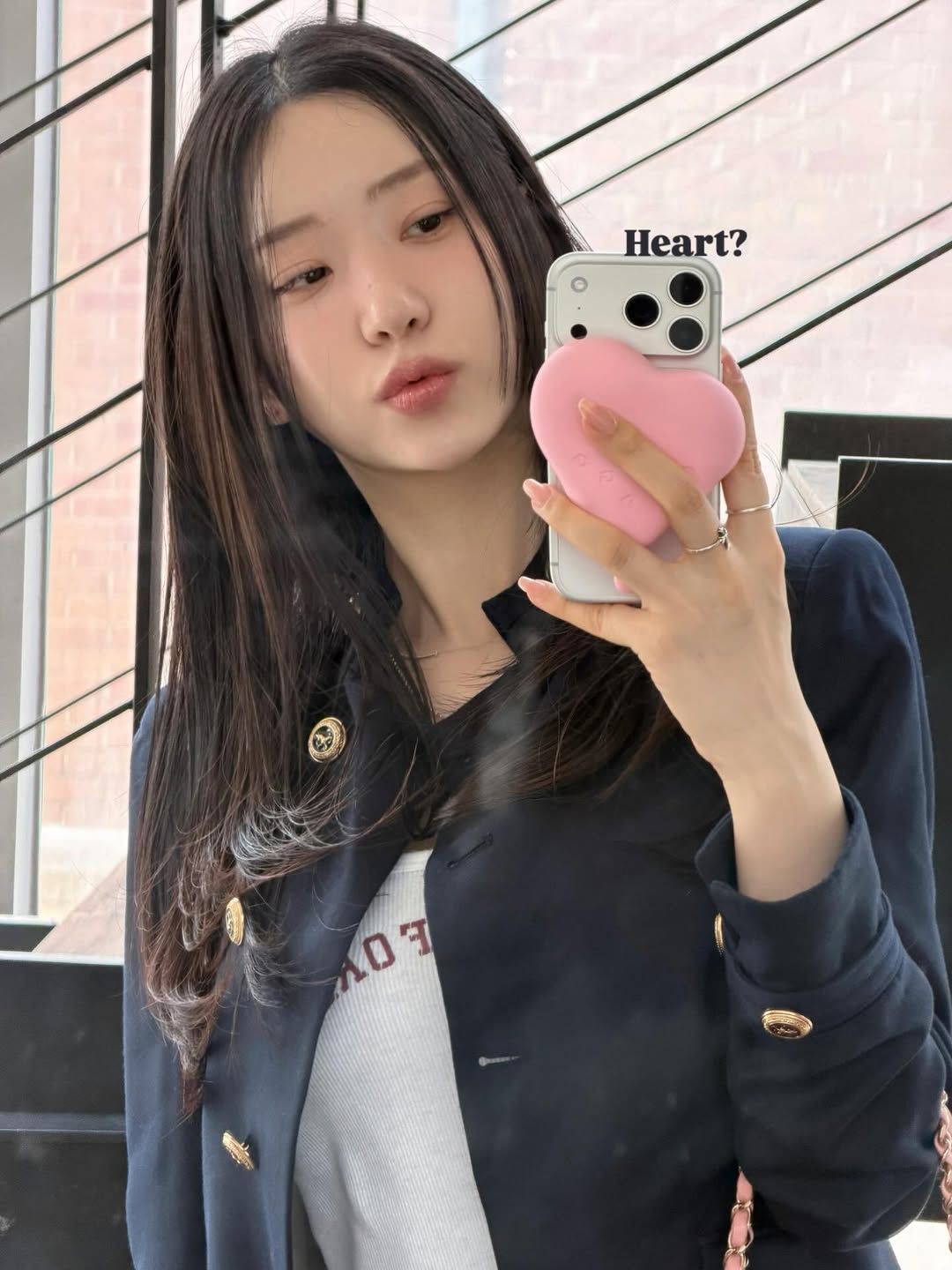一位女性在鏡子前自拍，穿著深色外套，手持一個粉紅色心形手機殼，臉上帶著唸嘴表情。