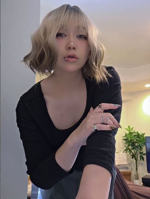 一位年輕女子，短髮呈波浪型，穿著黑色上衣，手上戴著多個銀色戒指，微微傾身面向鏡頭，背景為室內環境。