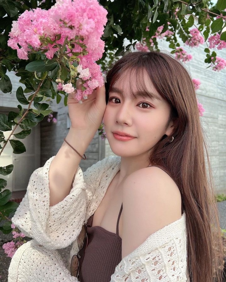 一名年輕女性在盛開的粉紅色花朵旁微笑，穿著輕薄的編織外套，背景模糊的建築物隱約可見。