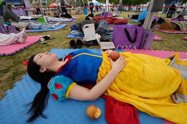 一名女性穿著白雪公主的服裝，躺在戶外草地上，身旁有幾個蘋果，背景中有其他人和一些物品。