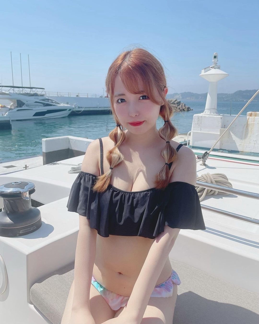 一位女孩穿著黑色露肩比基尼上衣，坐在遊艇上，背景是平靜的海面和幾艘船隻。