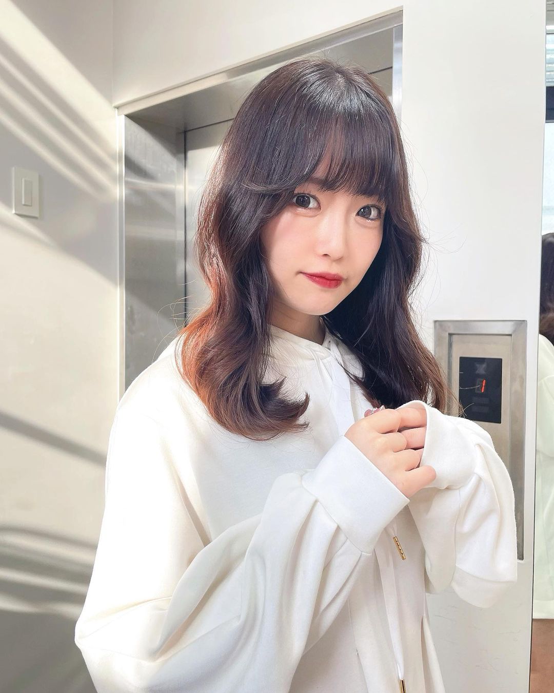 一位穿著白色衣服的女性在鏡子前微笑，背景是電梯的金屬門，柔和的光影效果增強了畫面的美感。