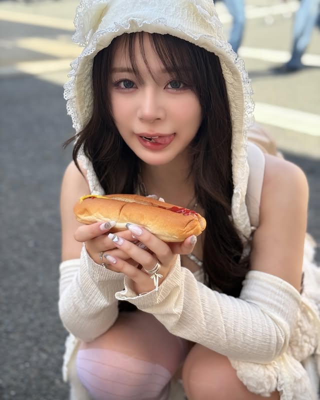 一位年輕女性穿著可愛的白色服裝，手持熱狗，微笑並舔著嘴唇，背景為戶外街道。